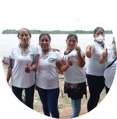 Four women stand in front of a river, smiling and giving thumbs-up. They are wearing white shirts and dark pants, with one woman wearing a face mask. The background includes boats and lush greenery across the river.