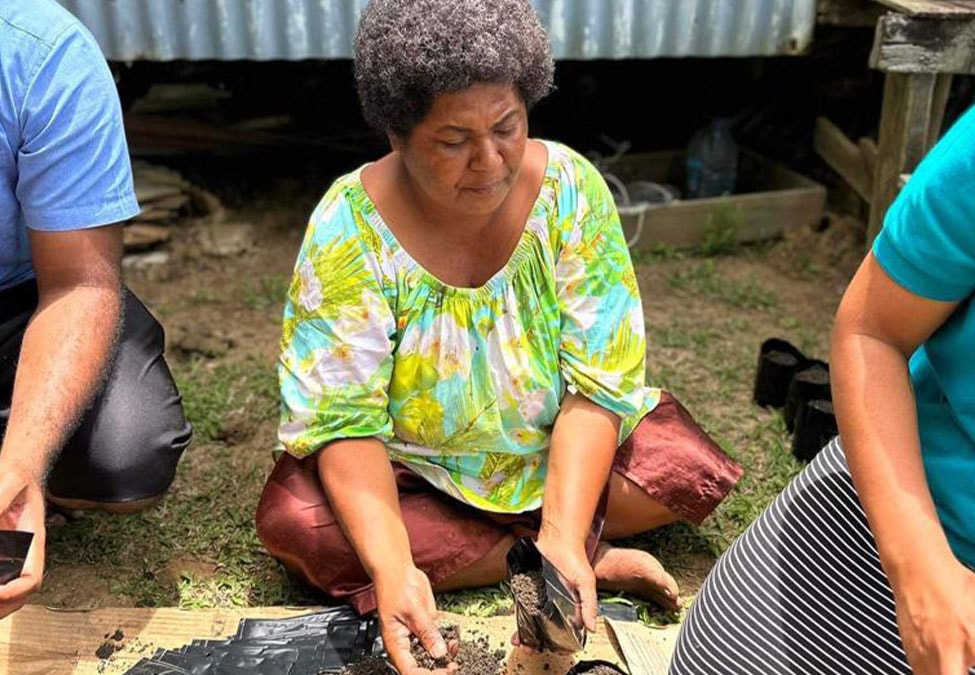 A woman with short curly hair is seated on the ground, wearing a colorful floral blouse and maroon pants. She is planting seedlings in small pots, with people on either side of her participating in the activity. They are outdoors, near a corrugated metal structure.