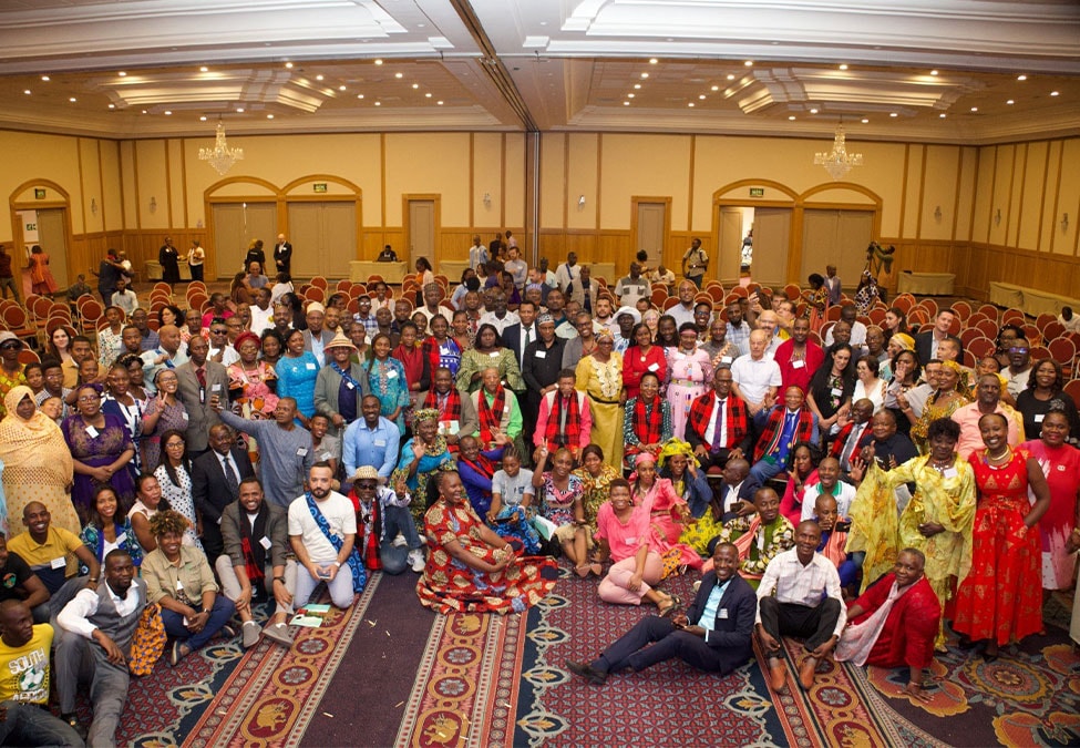 A large, diverse group of people, including men and women, are gathered in a hotel conference room with rows of chairs. Some are dressed in colorful traditional attire, while others wear casual or business attire. The room is adorned with chandeliers and high ceilings.