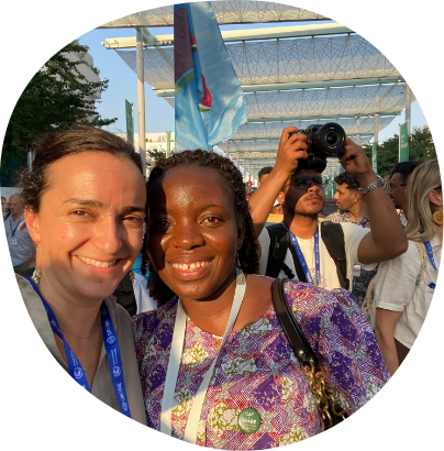 Two women are smiling for the camera in an outdoor setting with lanyards around their necks, suggesting they are at an event or conference. In the background, several people are also seen, one holding a professional camera. Trees and a canopy structure are visible above.