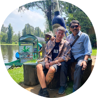 Two men are smiling and sitting outdoors on tree stumps near a lakeshore. The man on the left is wearing a patterned shirt over a black top, while the man on the right is in a light blue shirt and sunglasses. Behind them is a large bottle-shaped sculpture and greenery.