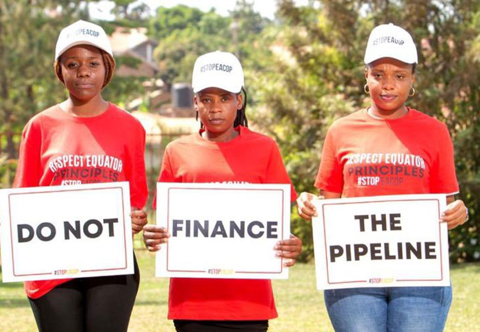 Three people are standing outdoors in a line, wearing white caps and red T-shirts that read "RESPECT EQUATOR PRINCIPLES STOP" followed by a hashtag. They each hold signs reading "DO NOT," "FINANCE," and "THE PIPELINE." Trees and a building are in the background.