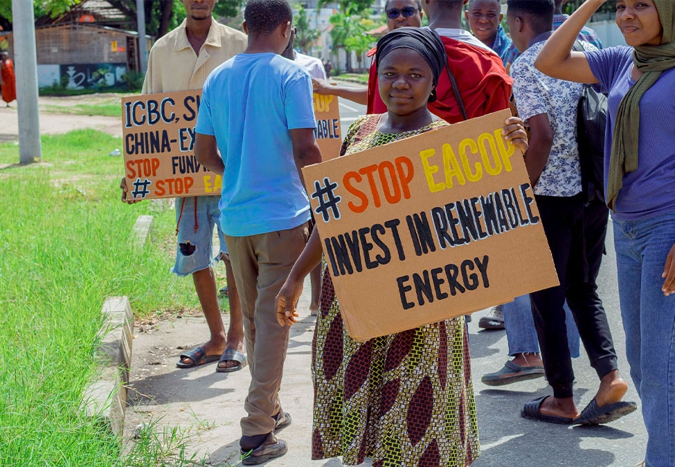 A group of people are participating in a protest. The focal point is a woman holding a cardboard sign that reads "#STOP EACOF INVEST IN RENEWABLE ENERGY." Others are also holding signs with similar messages advocating for environmental issues.